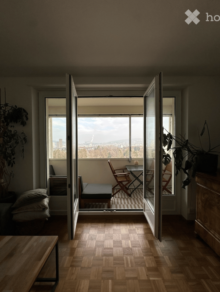 Modern living room with wooden flooring, large windows offering a city view, and a small seating area with a sofa, table, and chairs.