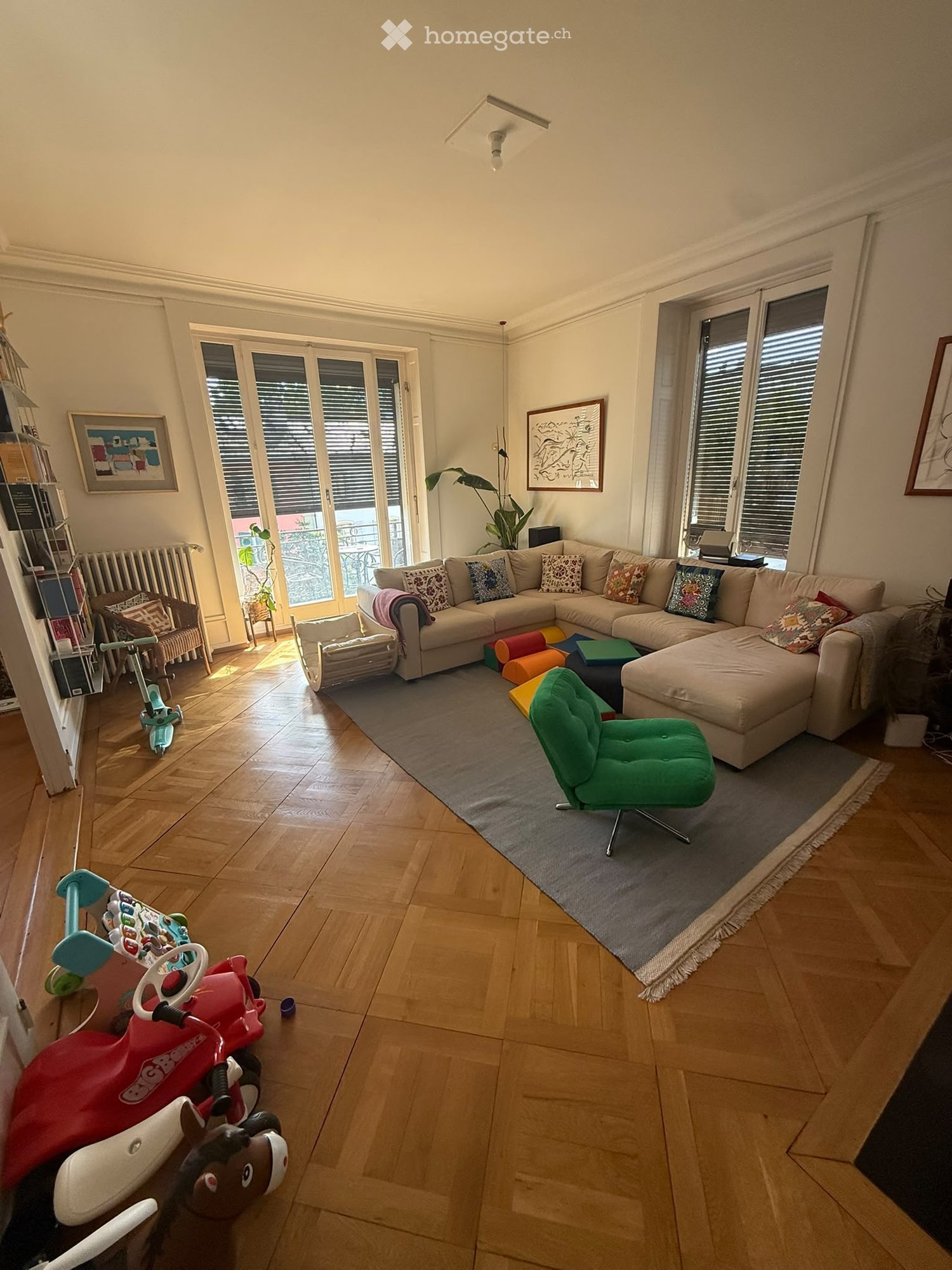 Bright living room with wooden flooring, large windows, a sectional sofa, a green armchair, and various toys including a toy car and a toy horse. There are also decorative pillows, a small table, and framed artwork on the walls.