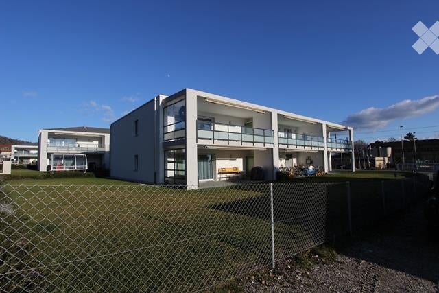 Modern two-story building with white exterior, multiple windows, and balconies. The building is surrounded by a fenced area with grass and gravel.