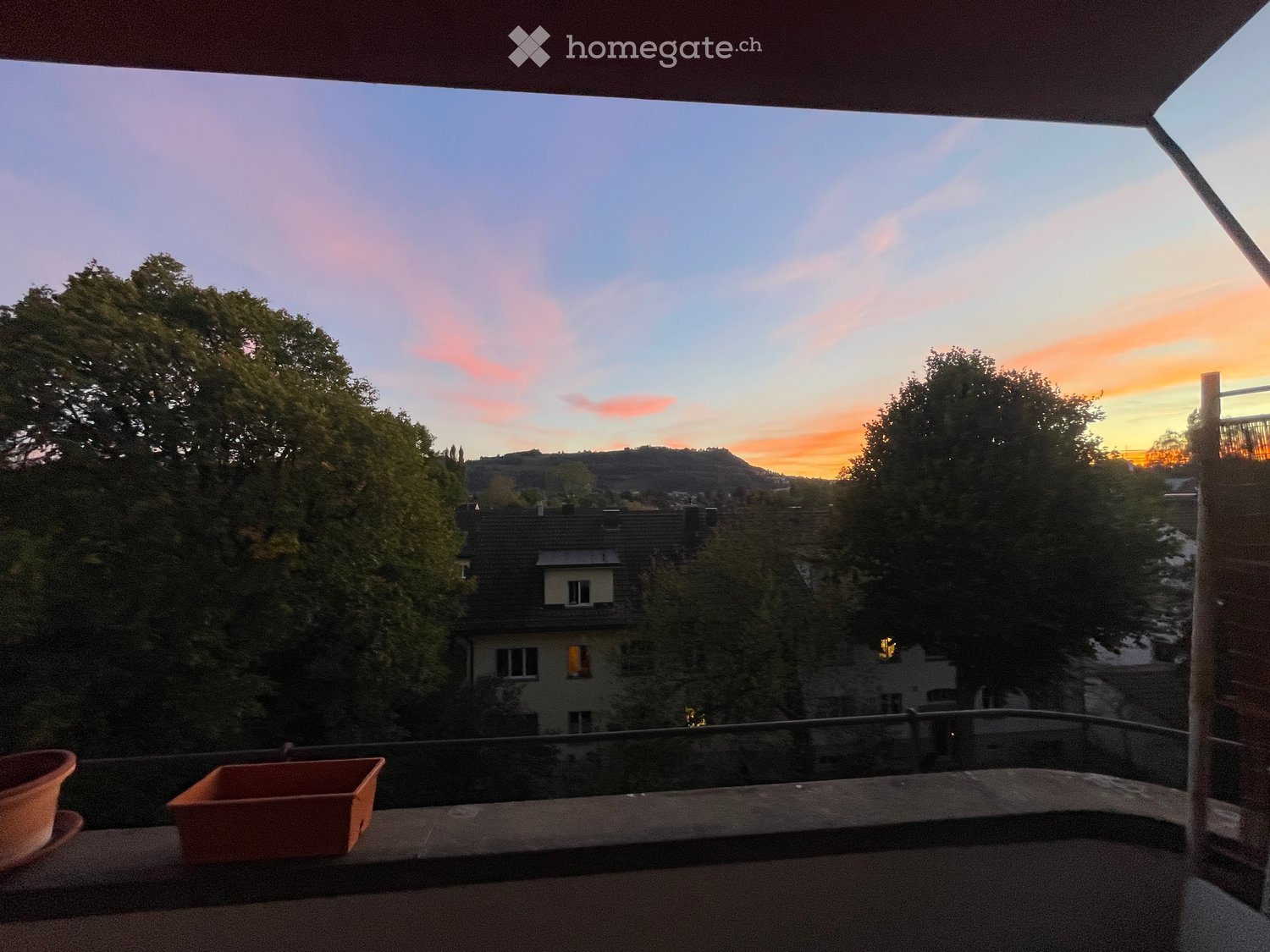 Balcony with a view of a cityscape at sunset, featuring trees and rooftops in the distance.