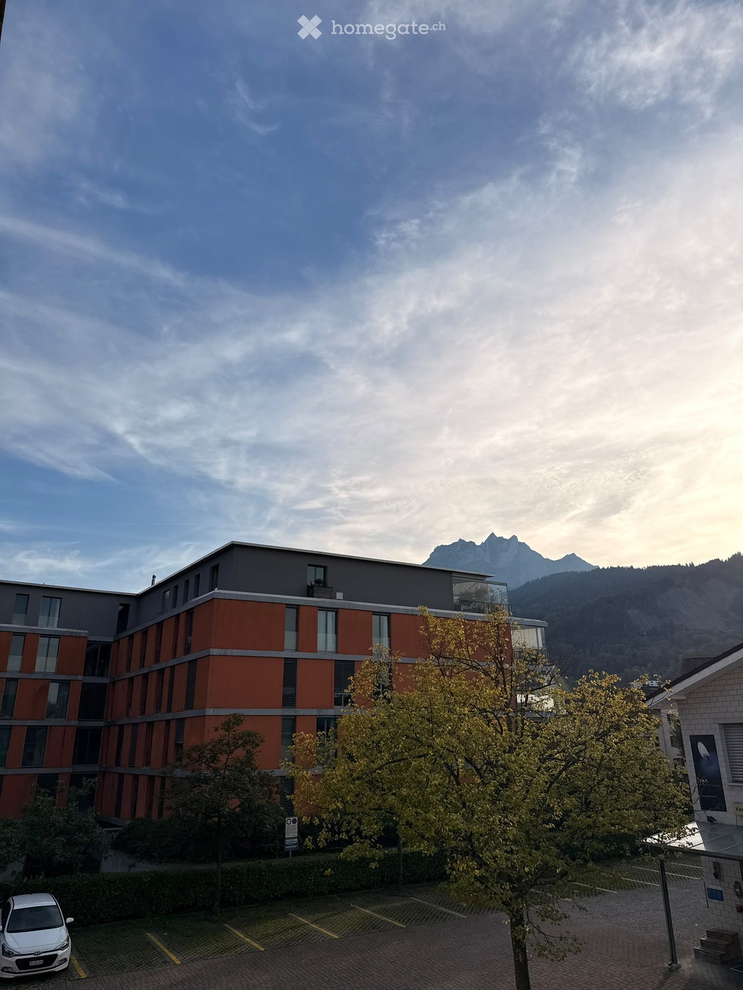Modern multi-story building with orange and gray facade, large windows, and a parking area in front, with trees and a scenic mountain view in the background.