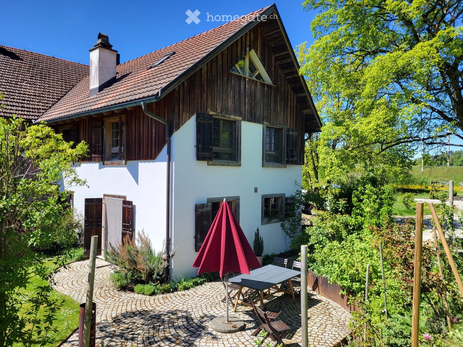 A charming two-story house with a red-tiled roof and wooden accents, surrounded by lush greenery. A patio area features a table, chairs, and an umbrella, creating a cozy outdoor space.