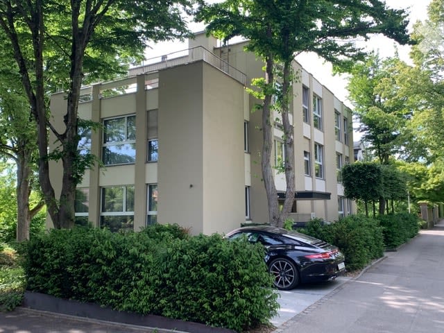 Modern multi-story building with large windows, surrounded by trees and greenery. A black sports car is parked in front.
