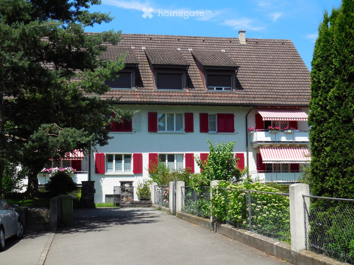 Two-story house with red shutters, white walls, tiled roof, and striped awnings on the balconies. The house is surrounded by greenery and has a small garden area with a fence.
