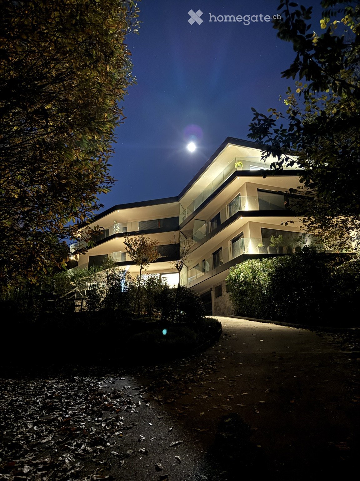 Modern two-story house with illuminated glass balconies and sleek architecture, surrounded by trees and shrubs.