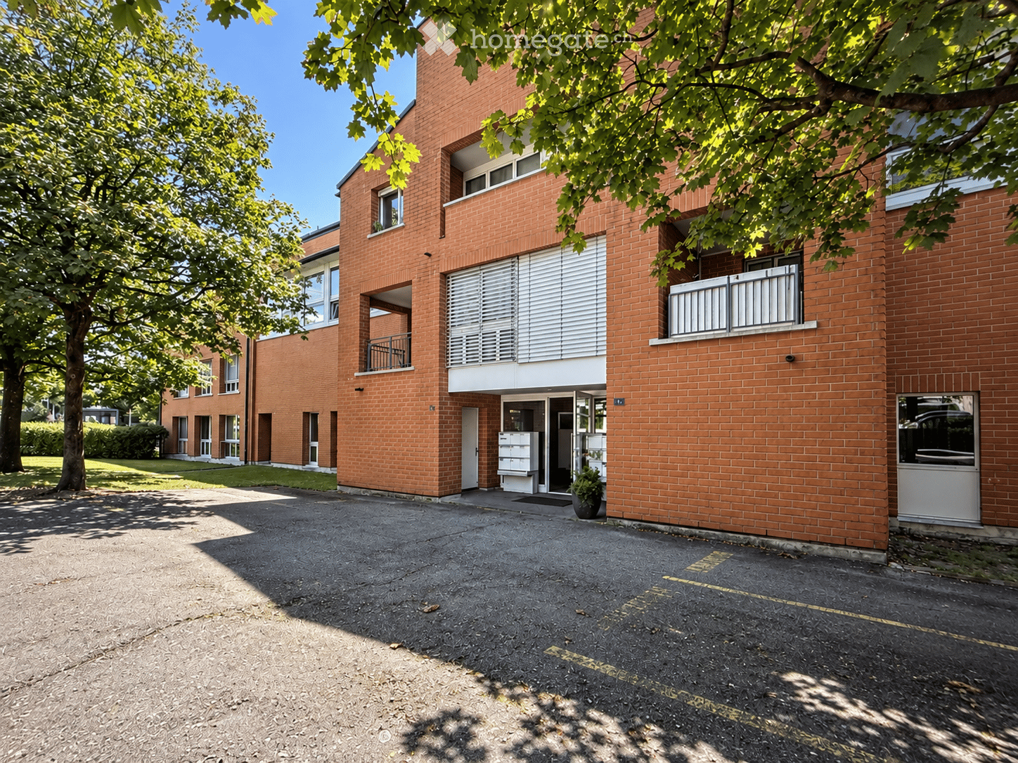 A multi-story brick building with balconies, large windows, and a paved parking area in front.