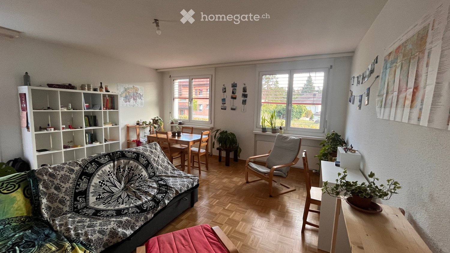 A cozy living room with a patterned sofa, wooden dining table, bookshelf, and potted plants. The room features large windows with a view of the outside and a mix of modern and rustic furniture.