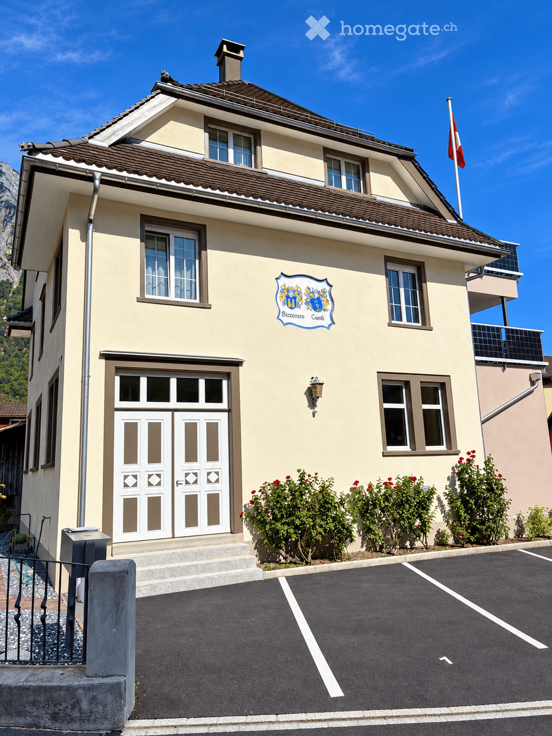 Two-story building with beige walls, multiple windows, and a brown roof. Features a double door entrance, a coat of arms sign, and a small garden area with flowering bushes. A parking lot is visible in front.