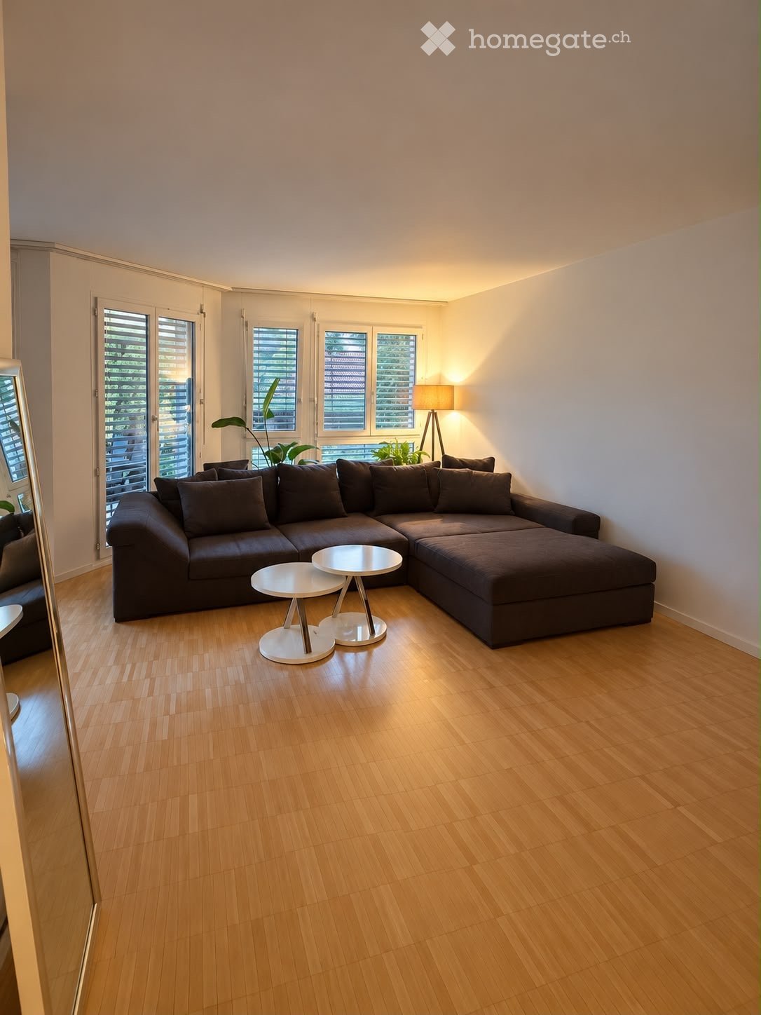 Modern living room with light wooden flooring, large sectional sofa, two white coffee tables, floor lamp, and indoor plants near the windows.