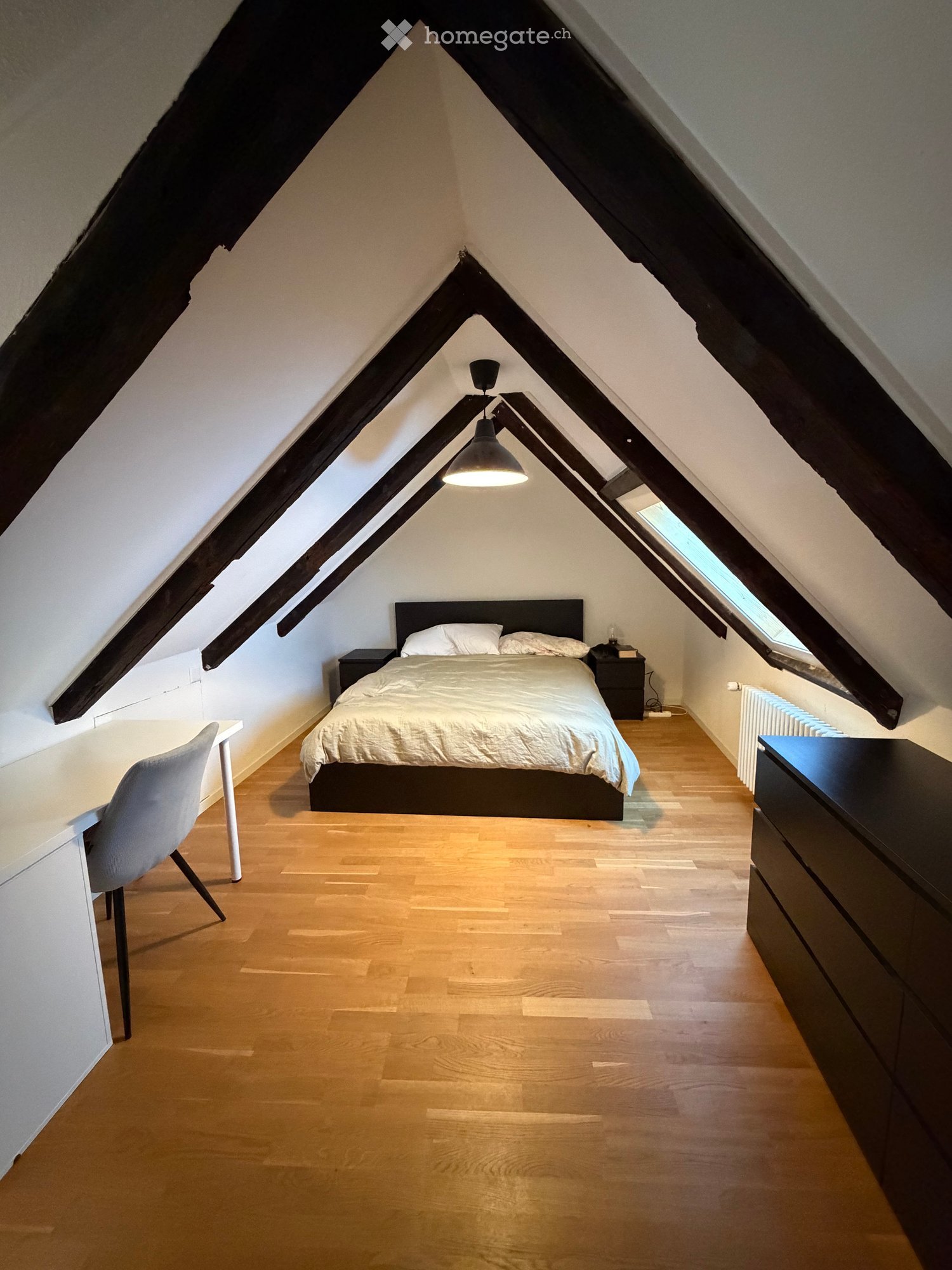 Cozy attic bedroom with wooden flooring, slanted wooden ceiling beams, and skylight windows. Features a bed with white bedding, a nightstand, a desk with a chair, and a hanging pendant light.