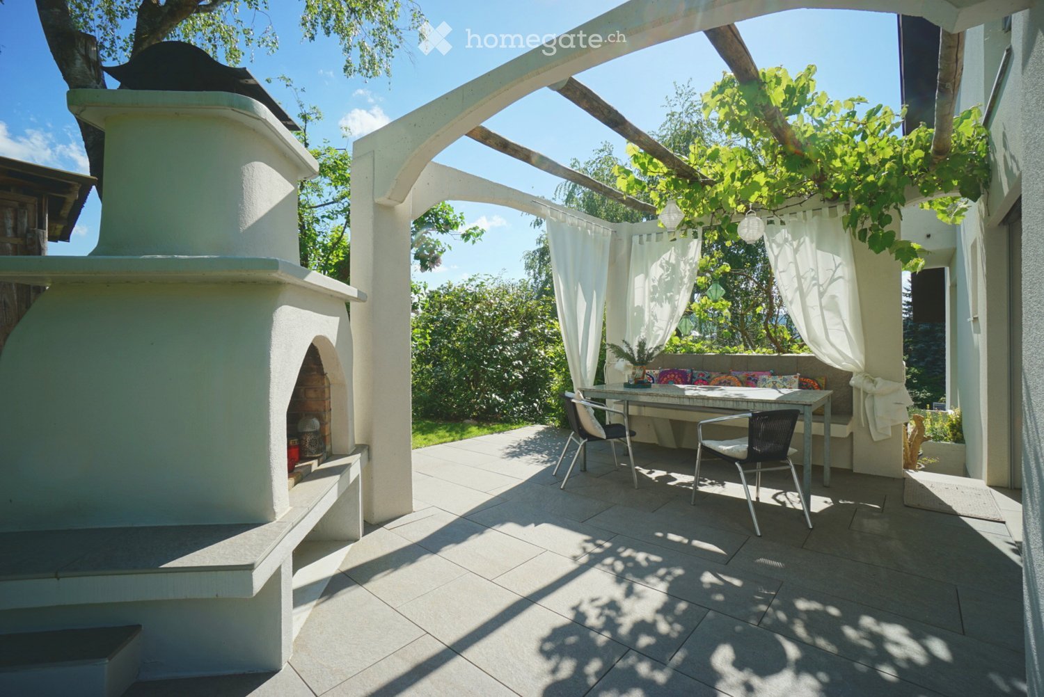 A covered outdoor patio with a stone floor, a white brick oven, a seating area with colorful cushions, a small table, and two chairs, surrounded by greenery and white curtains.