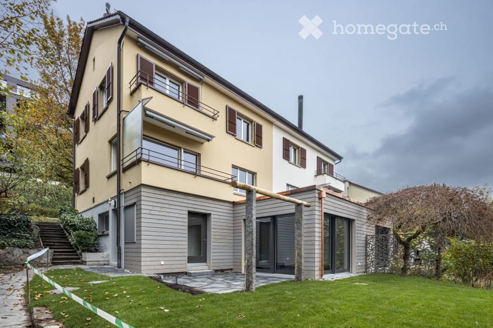 Two-story house with beige facade, multiple windows, balconies, and a modern extension with large glass doors.