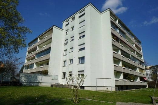 A multi-story residential building with white exterior walls, numerous windows, and balconies. The building is surrounded by a grassy area with a small tree and a playground visible in the background.