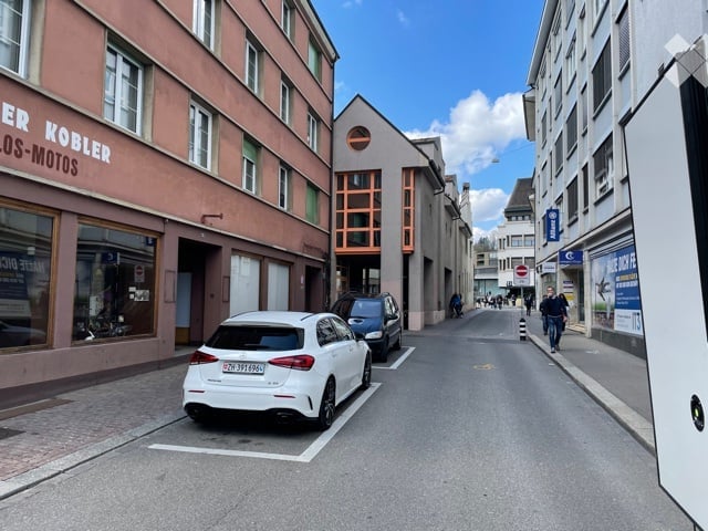 Urban street view with multi-story buildings, parked cars, and pedestrians. The buildings have a mix of modern and traditional architecture.