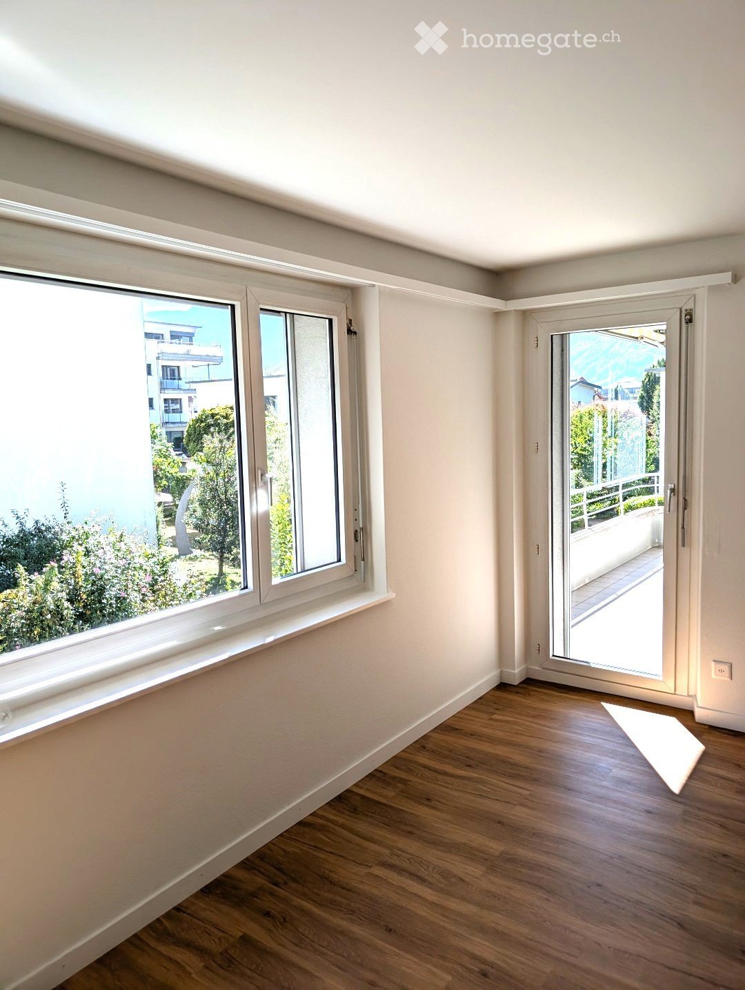 Empty room with wooden flooring, white walls, large windows, and a sliding glass door leading to a balcony.