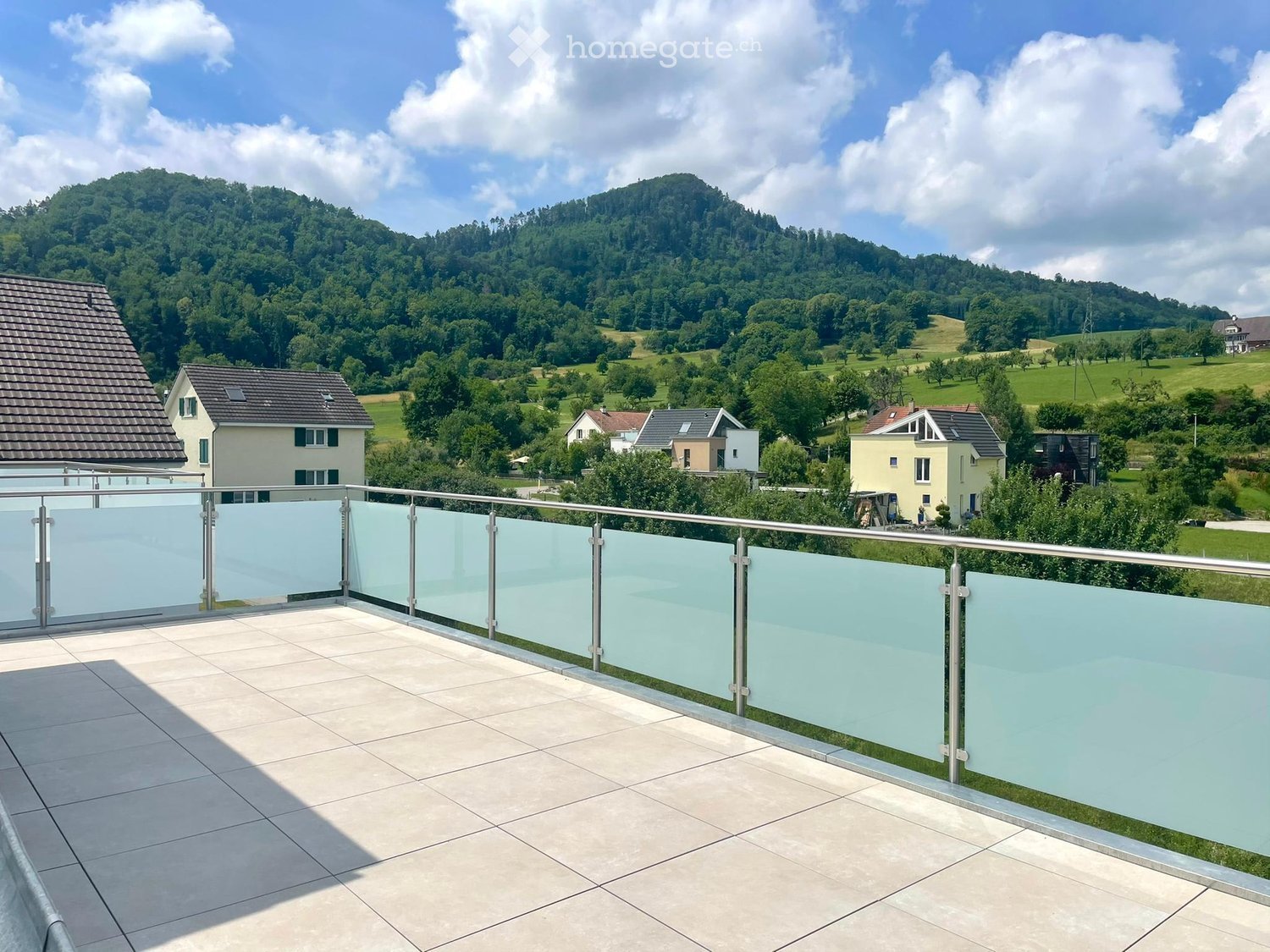 Balcony with modern glass railing, tiled floor, overlooking a green landscape with hills and houses.