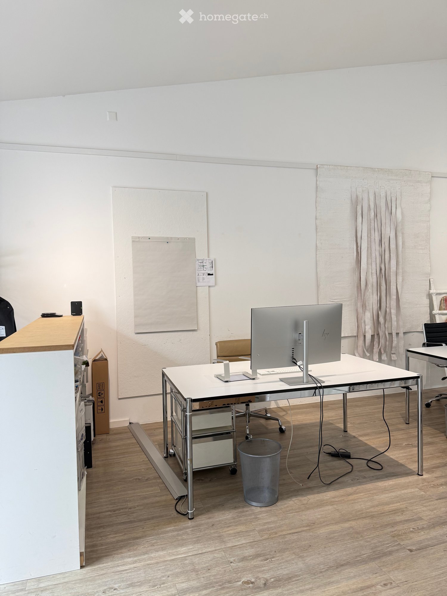 A modern, minimalist office space with a white desk, ergonomic chair, large screen, and a small trash can. The room features light wooden flooring and white walls.
