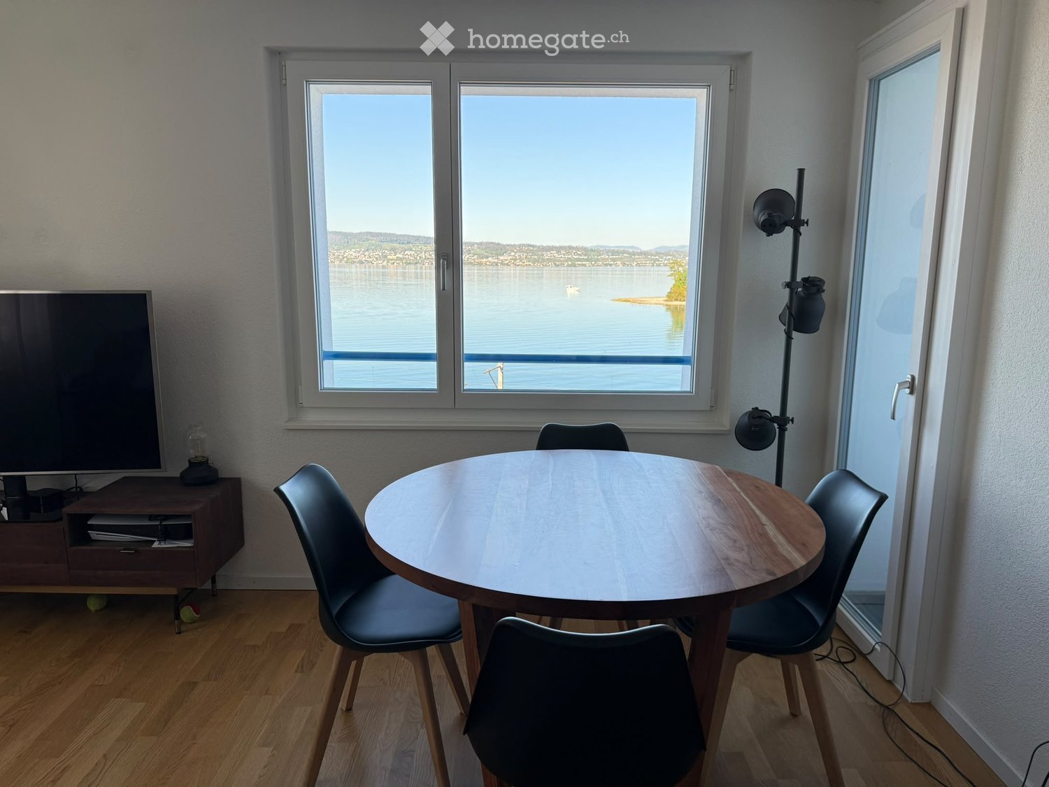 Bright room with wooden flooring, large windows overlooking a lake, round wooden table with four chairs, TV on a wooden stand, and a floor lamp.
