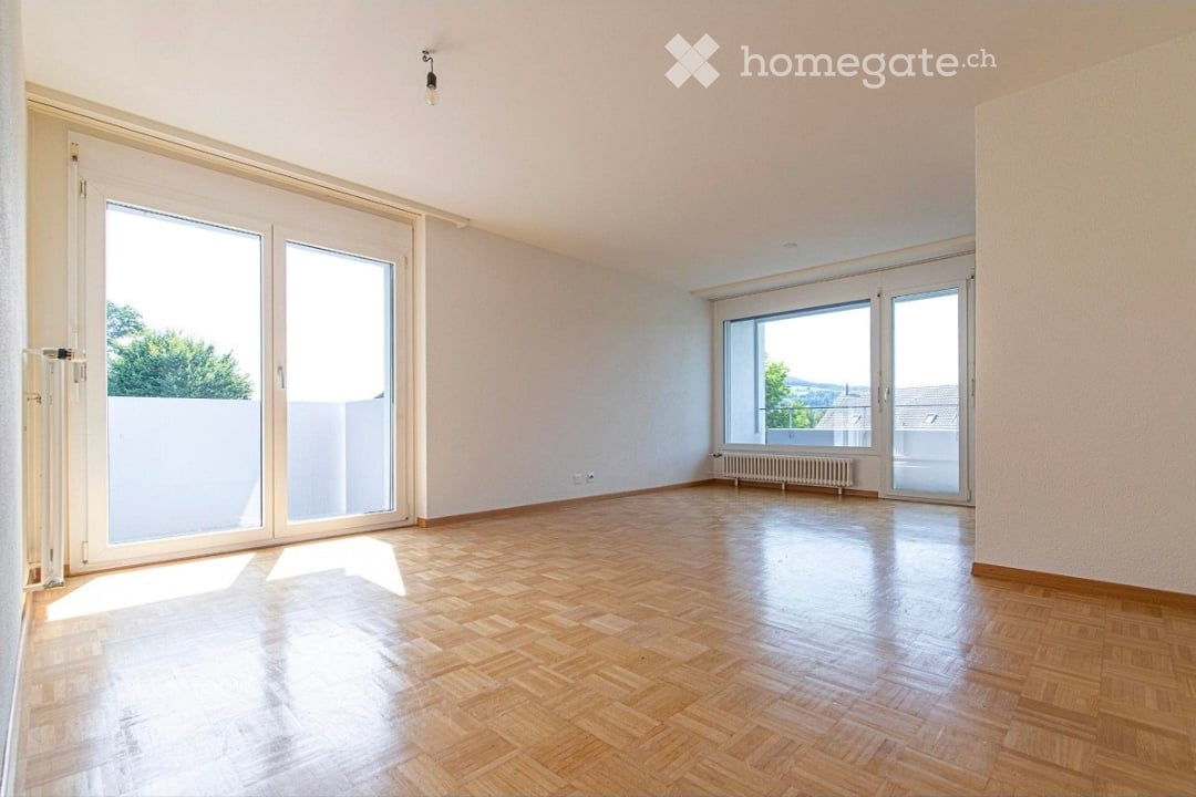 Empty room with wooden flooring, large windows, and sliding glass doors leading to a balcony.