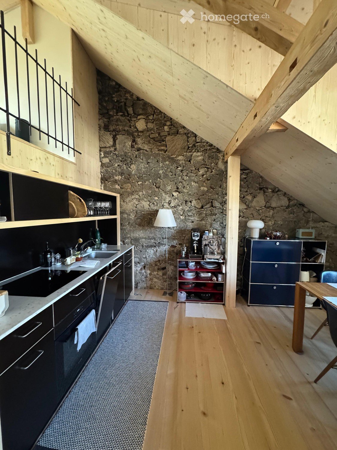 Modern kitchen with wooden floors, stone accent wall, black cabinetry, stainless steel appliances, and a small dining area.