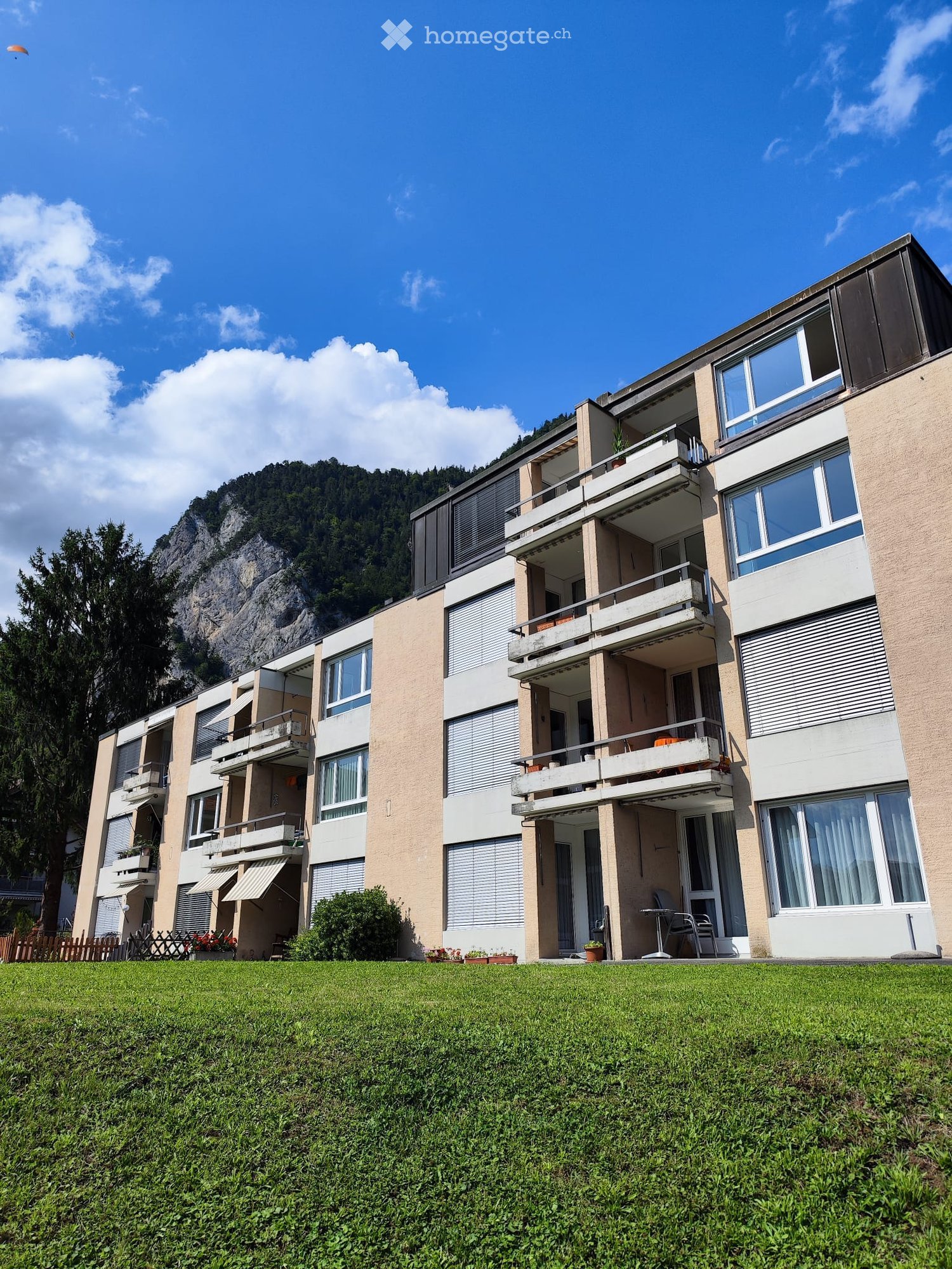Multi-story residential building with balconies, large windows, and a well-maintained lawn in front.