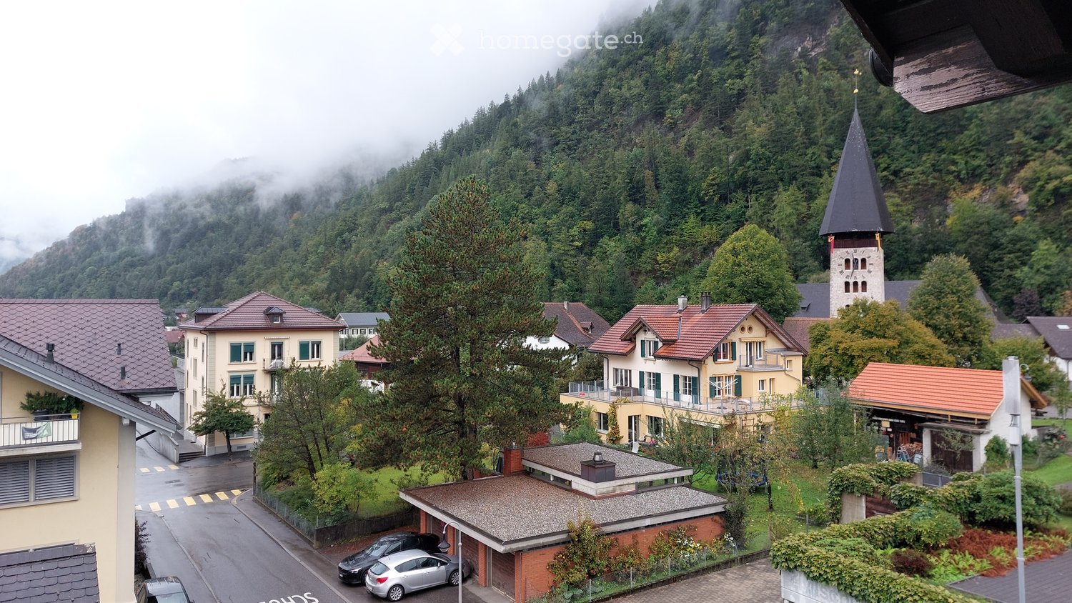 A scenic village view with traditional houses, a church tower, and surrounding forested mountains under a misty sky.