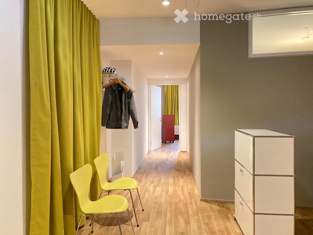 A narrow hallway with wooden flooring, two bright yellow chairs, a white wardrobe, and a coat hanging on a rack. A red suitcase is visible in the background.