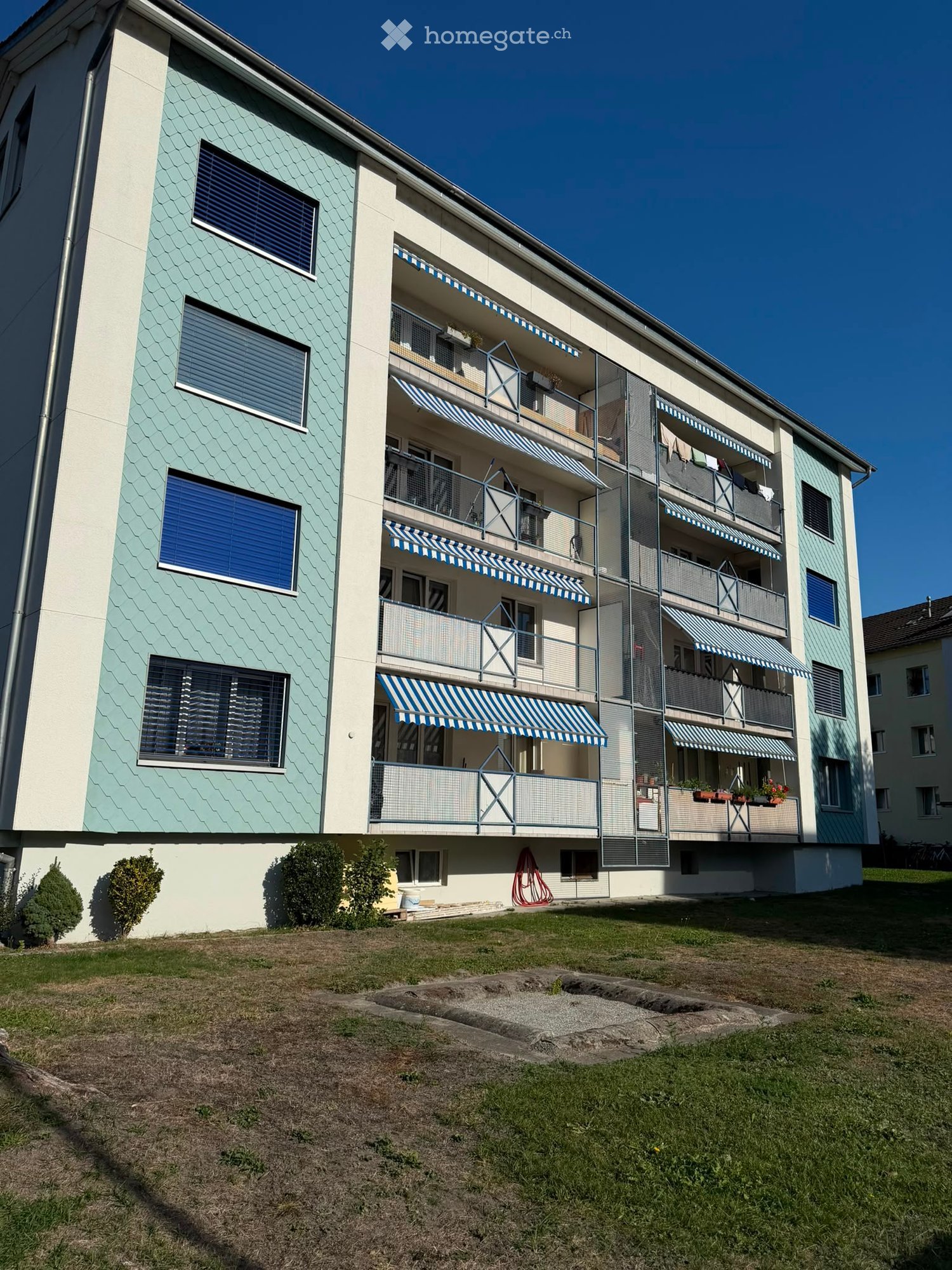 Multi-story residential building with light blue and white exterior, multiple balconies with striped awnings, and small garden area in front.