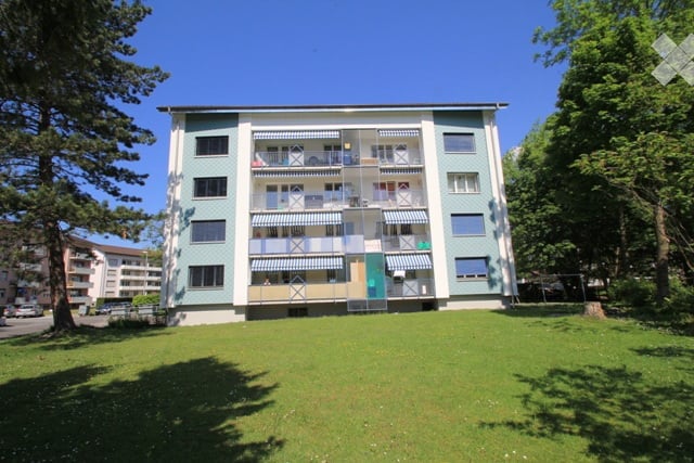 Multi-story apartment building with balconies, surrounded by trees and a grassy lawn.