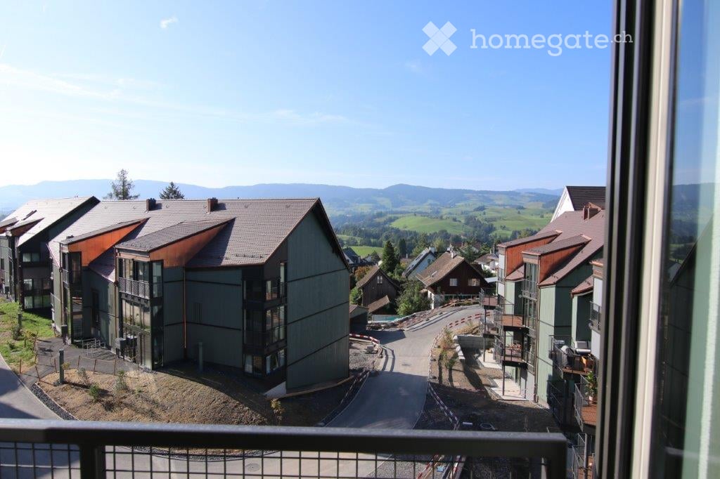 Balcony view overlooking a residential area with modern apartment buildings, green fields, and distant mountains under a clear blue sky.
