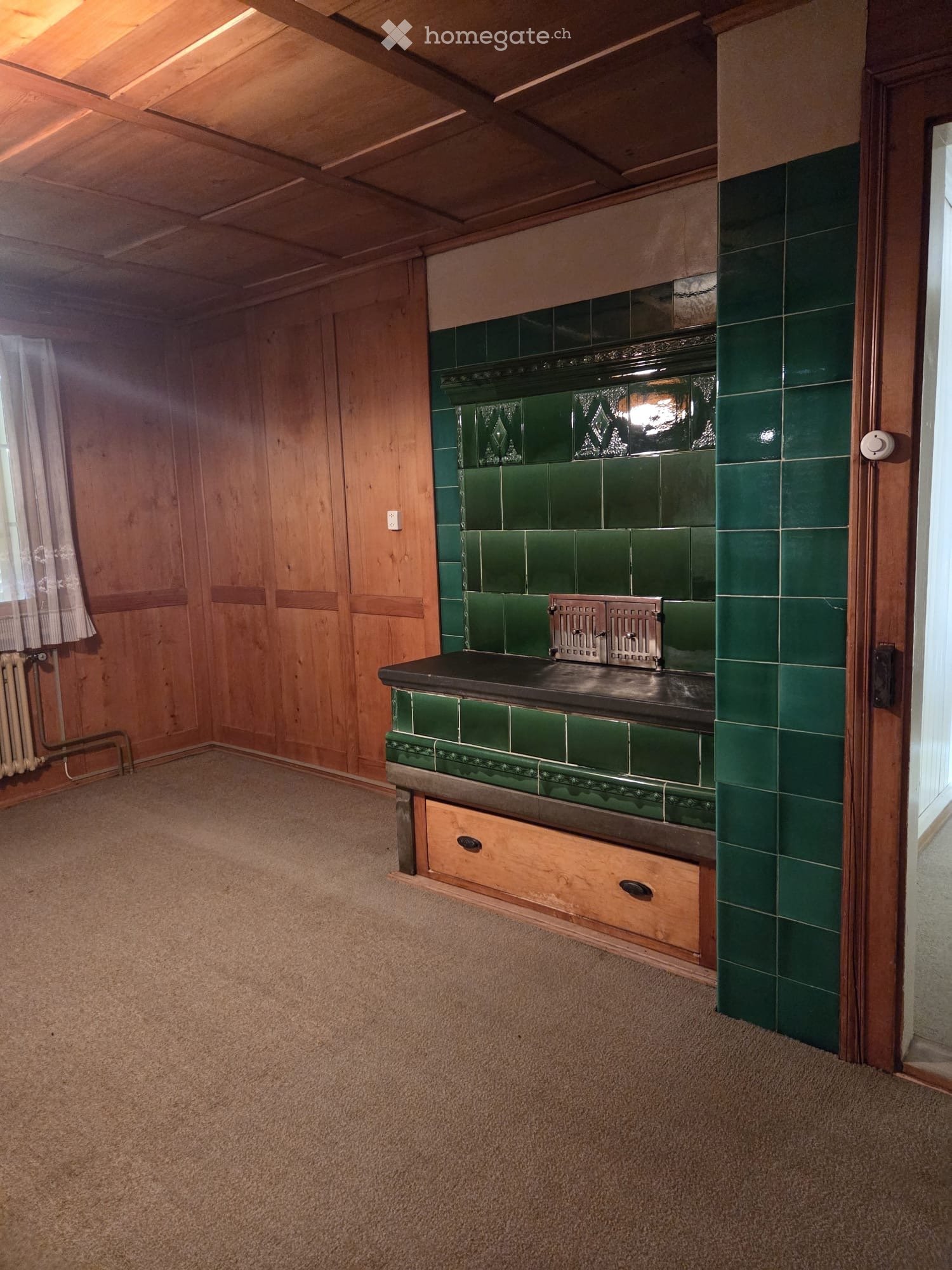 Empty room with wooden walls, green tiled stove, and carpeted floor.
