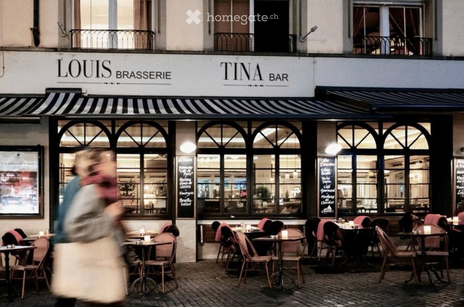 Outdoor seating area of a restaurant with tables and chairs, large windows showing interior, and a striped awning above.