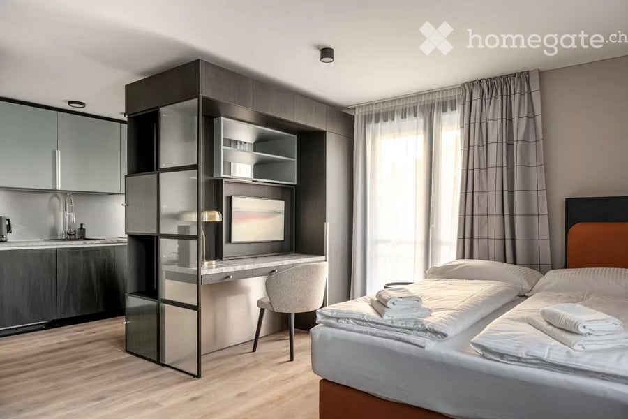 Modern, well-lit bedroom with a double bed, desk area, and built-in shelving unit. Features contemporary furniture and a sleek design.