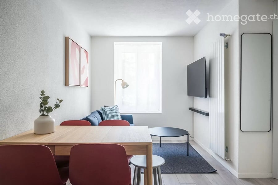 A modern, well-lit living room with a sofa, coffee table, dining table, chairs, a floor lamp, a wall-mounted TV, and a decorative plant on a side table.