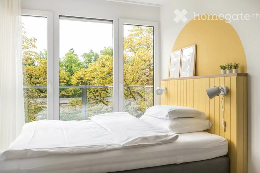 Modern bedroom with a large bed, white bedding, a wooden nightstand with a lamp, framed pictures, and small potted plants. Large windows offer a view of green trees outside.