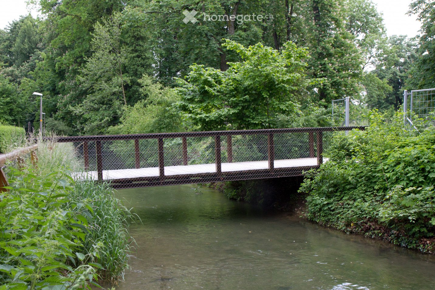 A small metal bridge with a chain-link fence spans a narrow stream surrounded by lush greenery and trees.