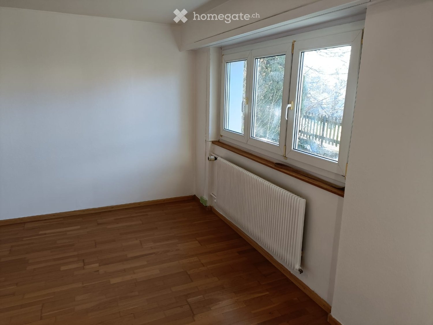 Empty room with wooden flooring, white walls, and a radiator under a large window with a view of a fenced outdoor area.