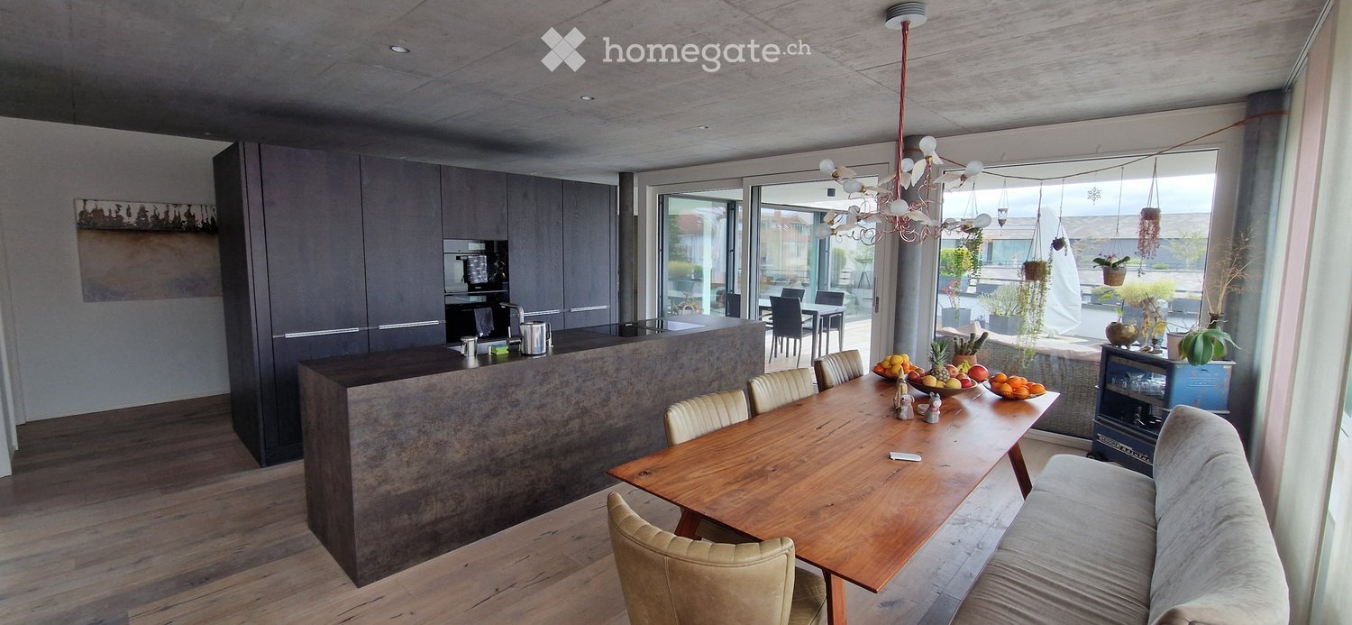 Modern open-plan living and dining area with a large wooden dining table, beige chairs, a grey sofa, and a kitchen island with cabinets and appliances.