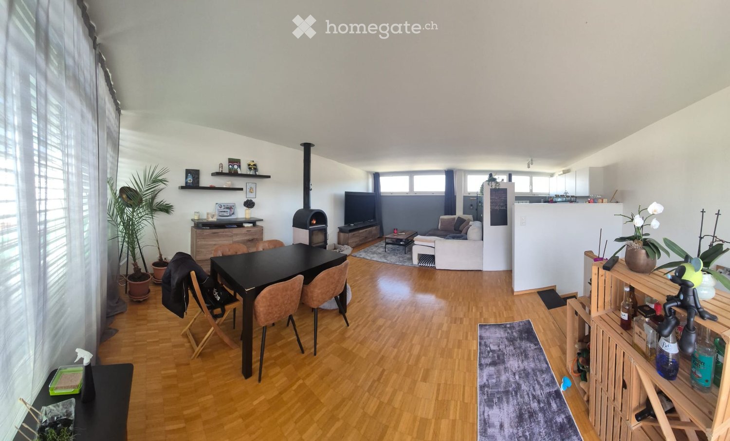 Open-plan living and dining area with wooden flooring, fireplace, sofa, coffee table, dining table with chairs, shelves, and plants.