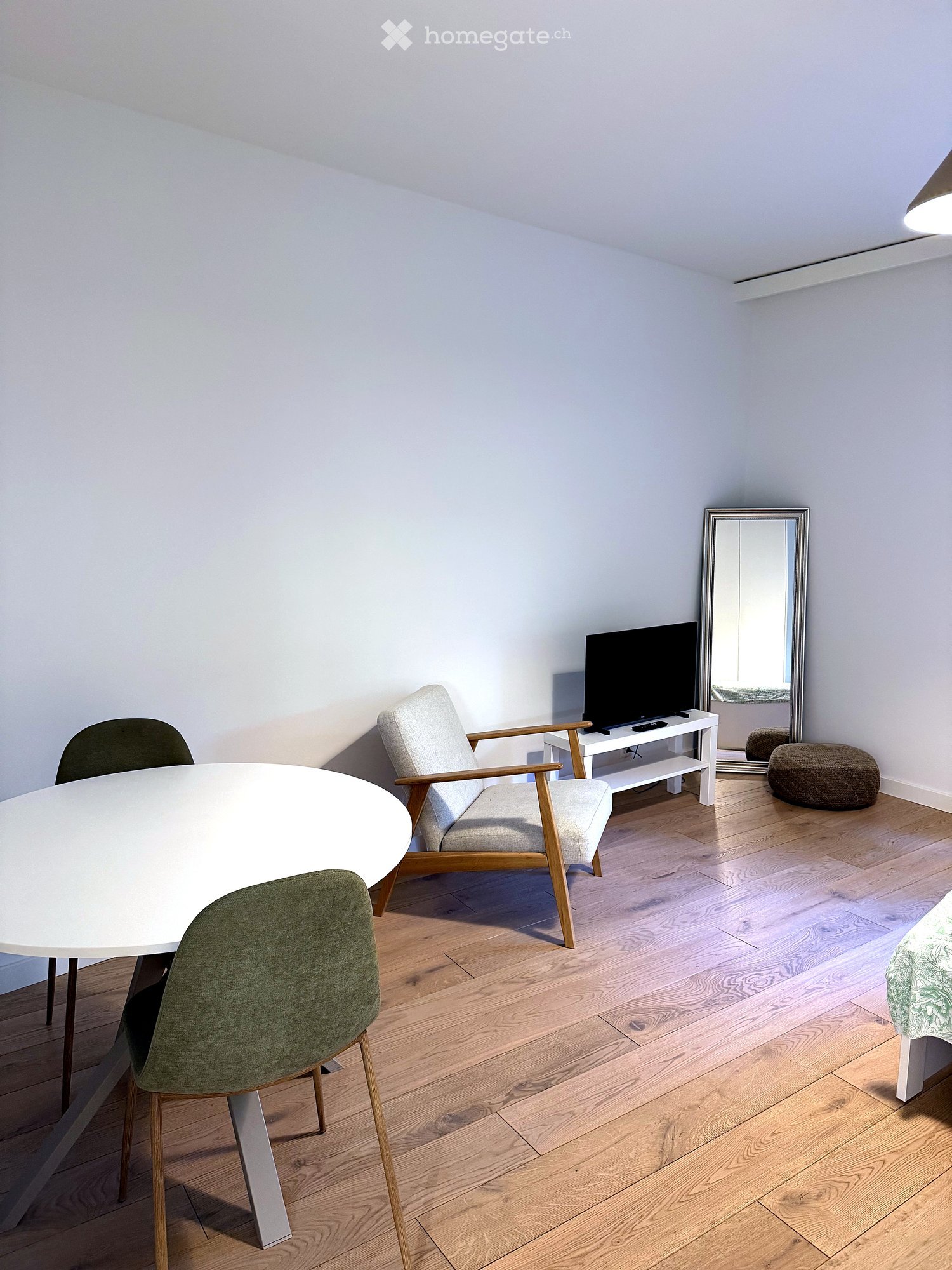 A modern, well-lit room with light wooden flooring, a white dining table with green chairs, a grey armchair, a television on a white stand, a round stool, and a large mirror on the wall.