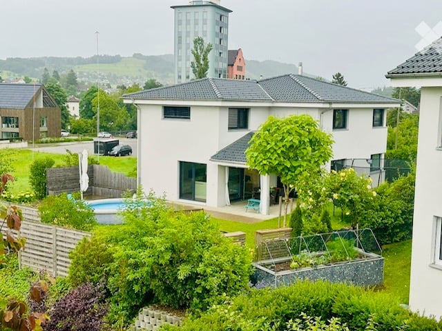 Modern two-story house with a grey roof, white walls, and large windows. Features a swimming pool in the garden, surrounded by greenery and a wooden fence.