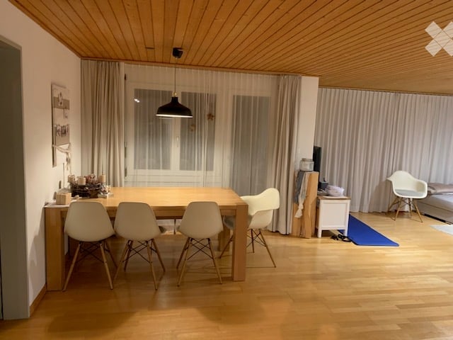 Dining room with wooden floor, dining table, four chairs, pendant lamp, and a TV screen in the background.