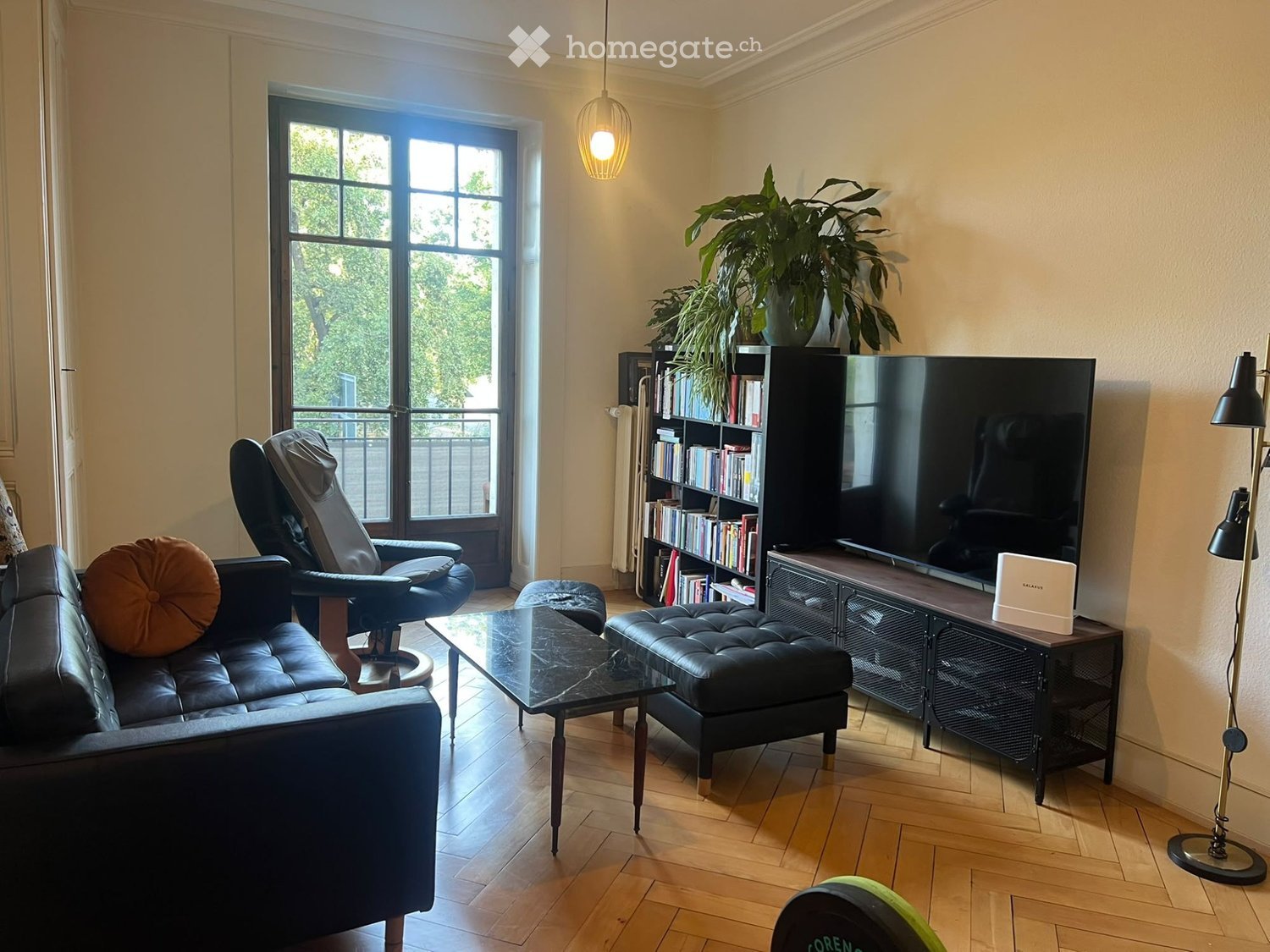 Modern living room with wooden flooring, large window leading to a balcony, a black leather sofa, a coffee table, an ottoman, a bookshelf, a television, and a floor lamp.