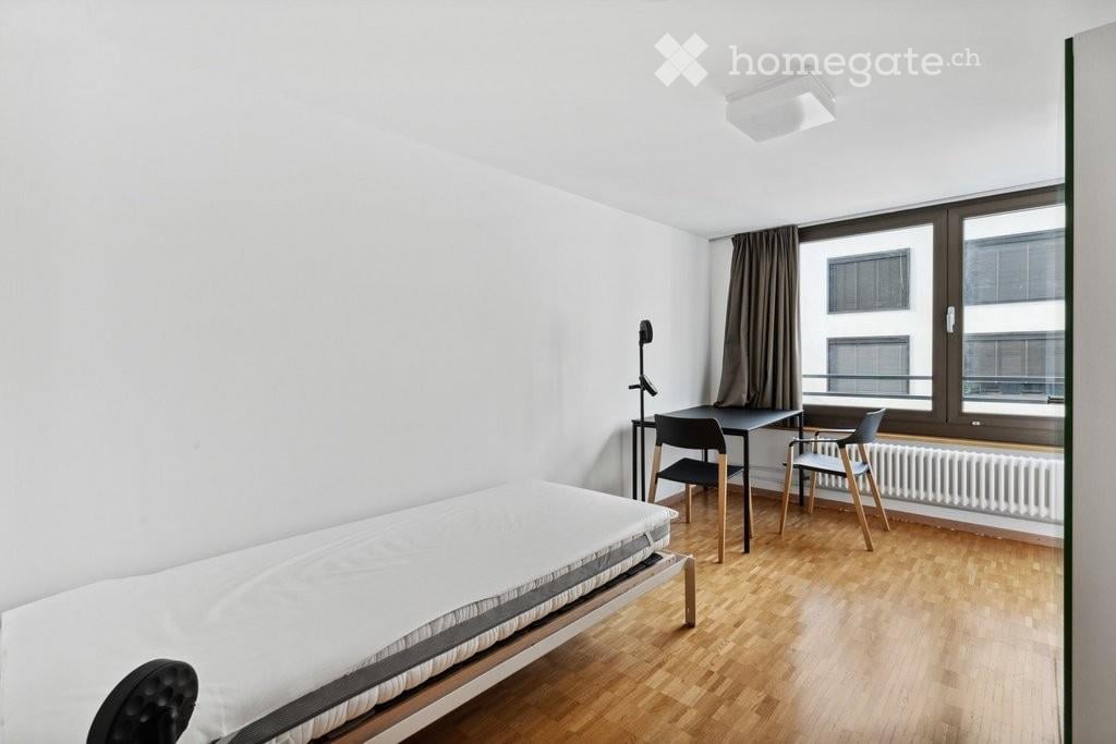 A small, modern bedroom with a single bed, a desk with a chair, and a window with curtains. The room features light-colored walls and wooden flooring.