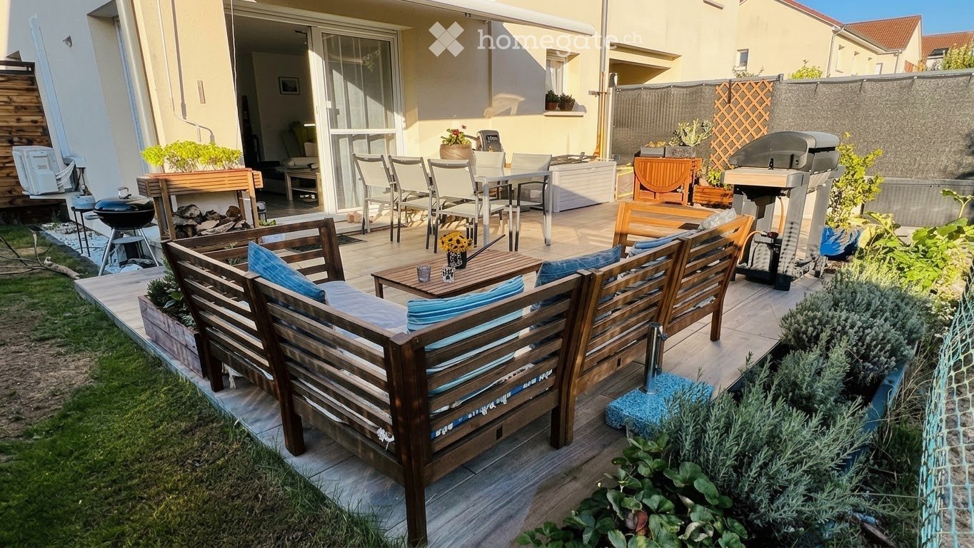 A well-maintained outdoor patio area with wooden furniture, including a sofa, chairs, and a dining set, surrounded by plants and a barbecue grill. The space is connected to an indoor living area through large glass doors.