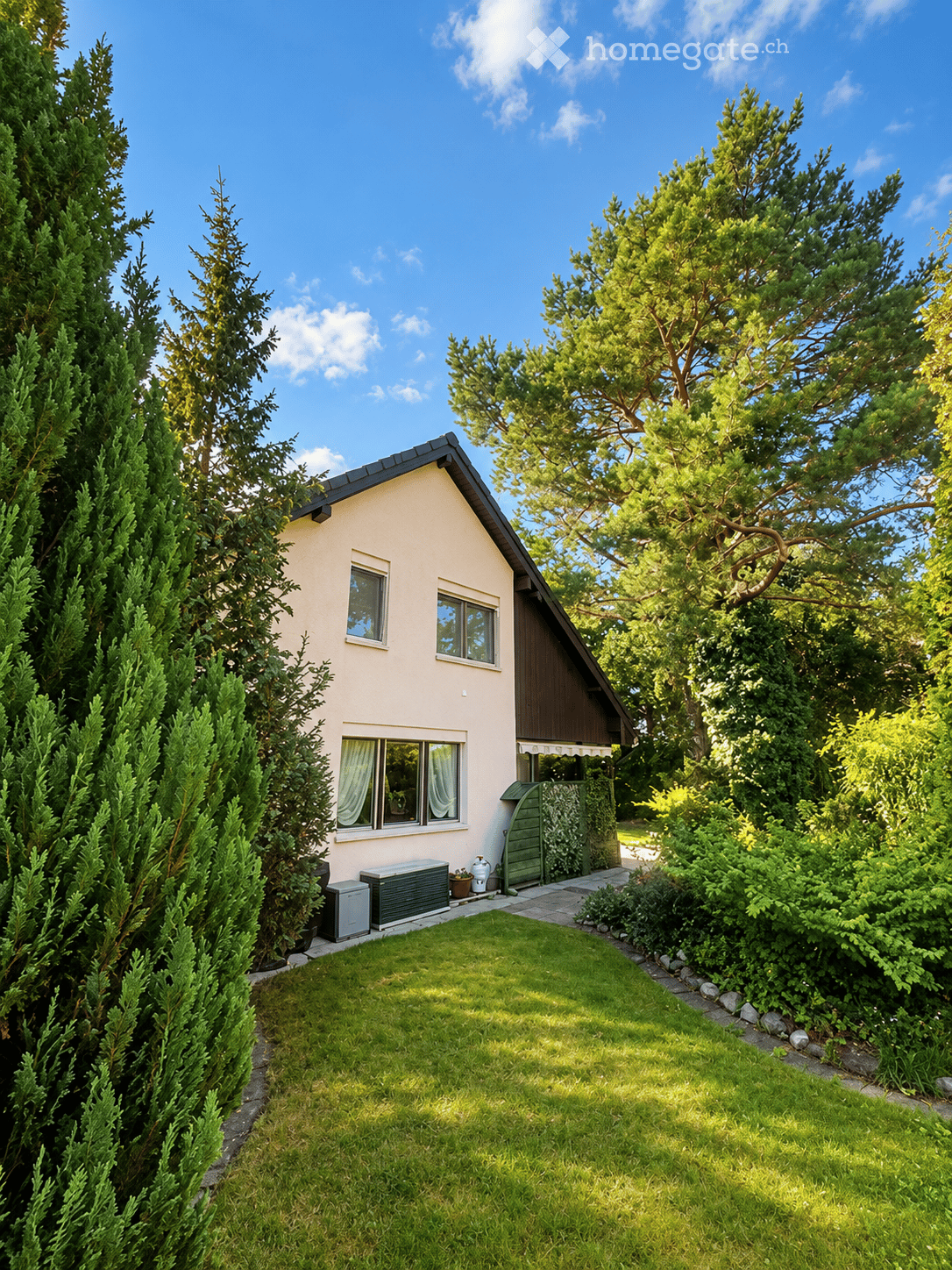 Single-story house with light beige walls, dark roof, and multiple windows. The house is surrounded by tall evergreen trees and has a small front yard with a neatly trimmed lawn and a small garden area near the entrance.
