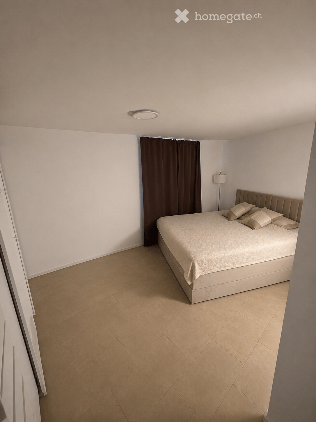 Modern bedroom with a large bed, beige pillows, a dark curtain, and a floor lamp near the window.