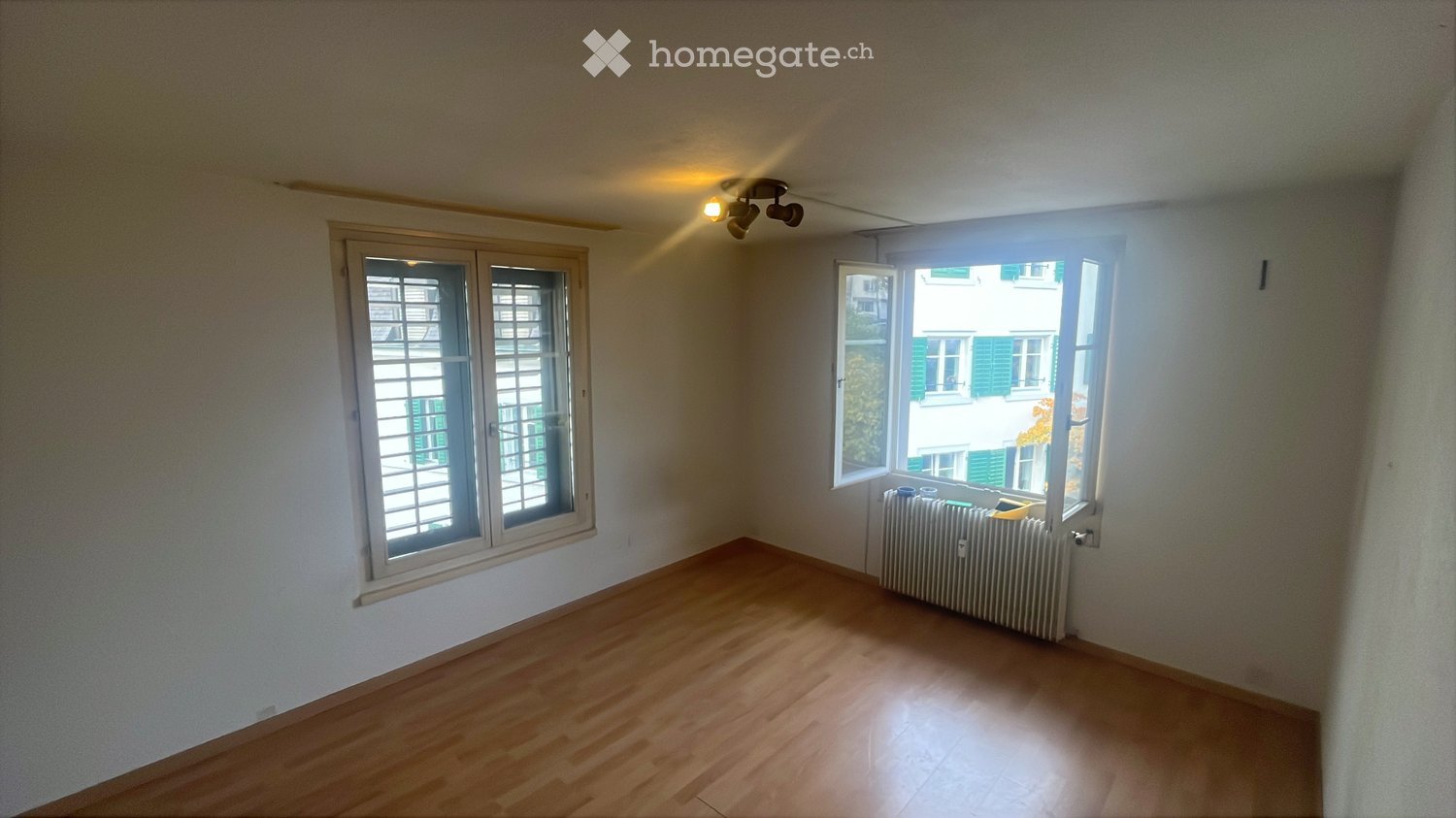 Empty room with wooden flooring, white walls, two windows, and a radiator under one window.