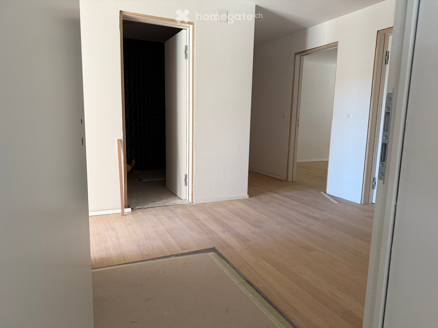 Empty room with light wooden flooring, white walls, and open doorways leading to other rooms.