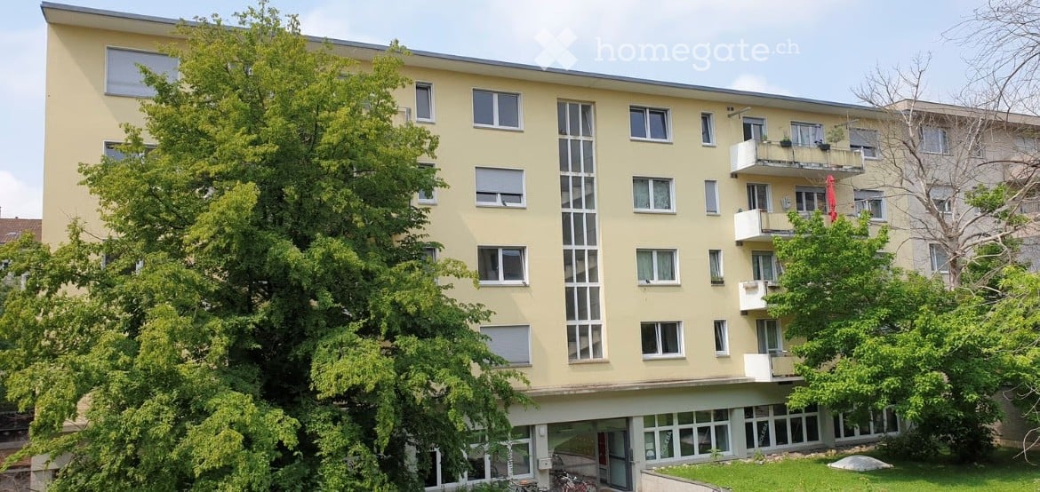 Four-story residential building with beige facade, multiple windows, and balconies on the upper floors.
